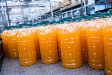 Das Bild zeigt eine Reihe großer Plastikflaschen, die mit orangener Flüssigkeit gefüllt sind und auf einem Förderband in einer Produktionshalle stehen.