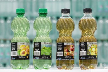 Das Bild zeigt vier PET-Flaschen mit unterschiedlichen grünen und schwarzen Verschlüssen und Etiketten, die verschiedene Fruchtsäfte darstellen, in einer Produktionsumgebung.