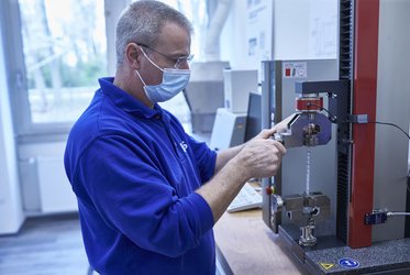 Ein Mann mit Schutzmaske und blauer Arbeitskleidung bedient ein Laborgerät zur Prüfung von Materialfestigkeit in einem hellen Raum.