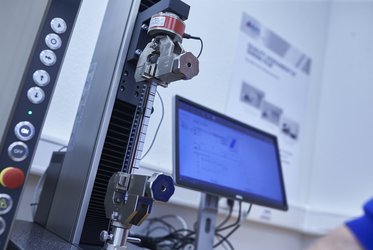 Ein Computerbildschirm zeigt Messergebnisse, während ein Prüfgerät mit einer eingespannten Probe im Vordergrund zu sehen ist.