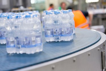 Das Bild zeigt mehrere verpackte Wasser-Sixpacks auf einem Förderband in einer hellen industriellen Umgebung.