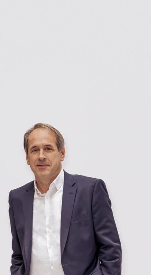 Das Bild zeigt den KHS-Kontakt Frank Fretwurst, einen Mann mittleren Alters in einem dunkelblauen Anzug und einem weißen Hemd, der vor einem schlichten, hellen Hintergrund steht und freundlich in die Kamera blickt.