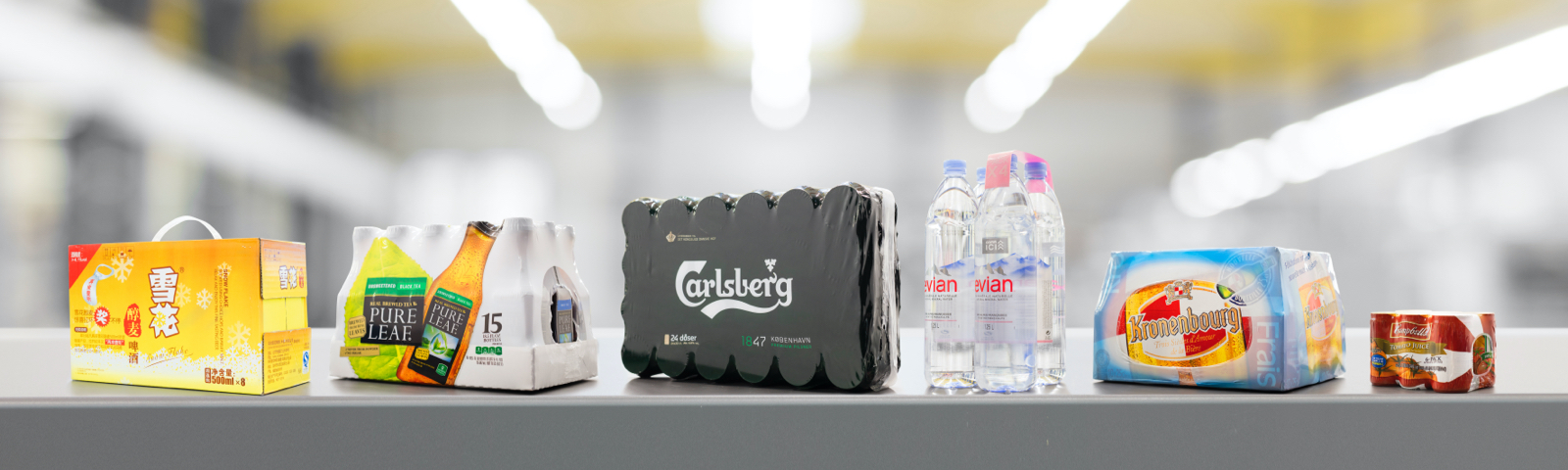 Das Bild zeigt verschiedene Getränkeumverpackungen, darunter Marken wie Carlsberg, Evian, Pure Leaf und Kronenbourg, die in unterschiedlichen Verpackungsarten nebeneinander aufgereiht sind, vor einem unscharfen industriellen Hintergrund.
