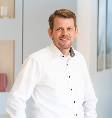 Das Bild zeigt Sebastian Wenderdel, einen lächelnden Mann mit kurzen, hellbraunen Haaren, der ein weißes Hemd trägt und in einer modernen, hellen Umgebung steht.